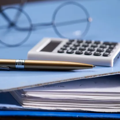 Black wire-rimmed glasses, white calculator and gold pen on light blue accounting ring-binder folder 